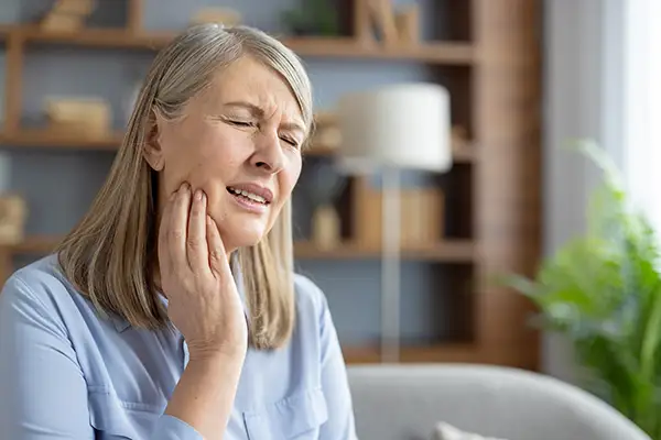Elderly woman sitting at home, holding her jaw and grimacing in pain from a cavity.