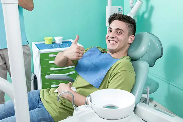 A grinning young man sitting in the dentist chair with a bib on and giving a thumbs up before his dental appointment.